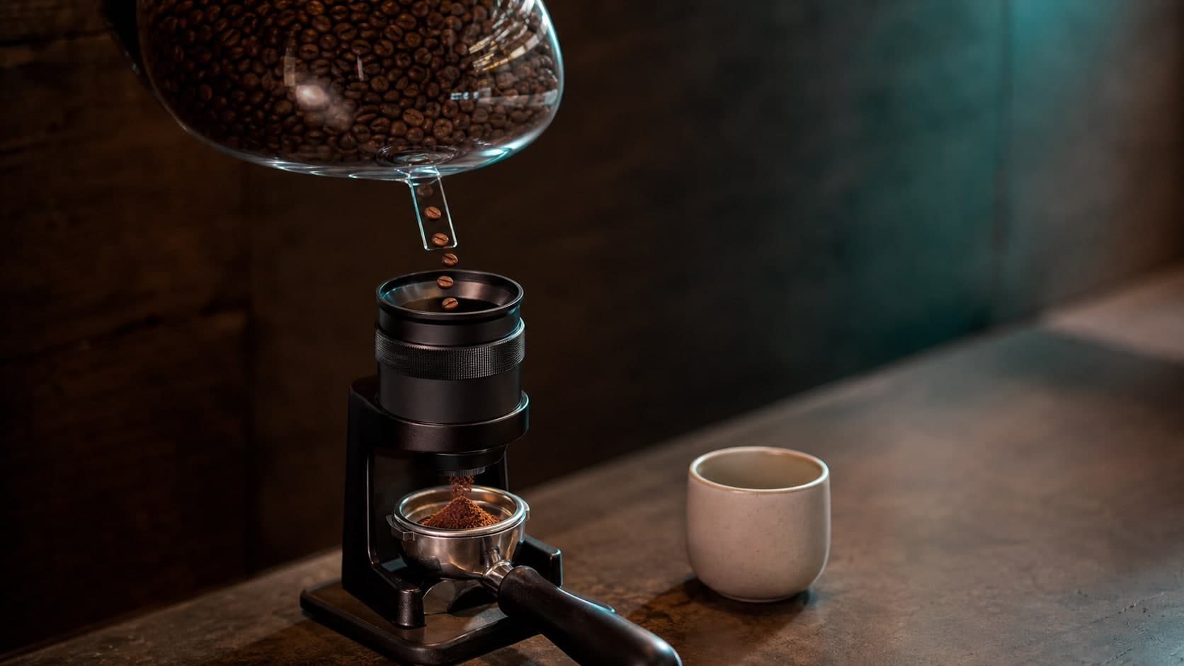 Coffee beans fall from a large glass hopper into a small grinder that drops grounds into a portafilter beside a ceramic cup.