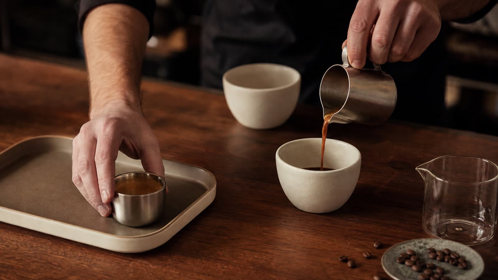 A barista sets a small metal cup in a tray while pouring espresso into a ceramic cup beside an empty cup and glass vessel.